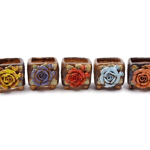 May include: Five small square ceramic flower pots with different colored flowers. The pots are brown with a textured surface and have three small feet on the bottom. The flowers are yellow, blue, red, light blue, and orange.