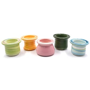 May include: Five small, colorful ceramic pots with a spool-like design. The pots are in shades of green, orange, pink, dark green, and blue, each with a contrasting rim. They are arranged on a white background.