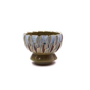 May include: A ceramic bowl with a pedestal base, featuring a textured exterior with vertical carved grooves. The upper portion is glazed in shades of light blue and white, while the lower base and interior are finished in a glossy olive green glaze.