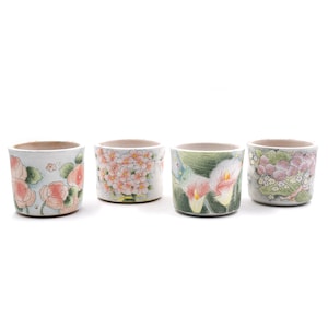 May include: Four small, ceramic cups with floral designs. Each cup features a different pattern: pink roses, peach blossoms, white calla lilies, and pink hydrangeas. The cups are cylindrical with a light-colored rim.