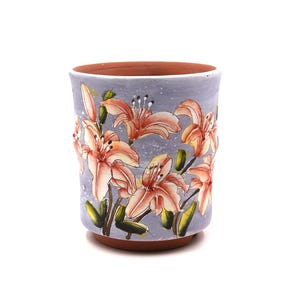 Può includere: Un vaso cilindrico in ceramica con interno e bordo in terracotta. L'esterno presenta un disegno dipinto a mano di gigli arancioni chiari con foglie verdi su uno sfondo blu pervinca maculato, caratterizzato da una finitura liscia.