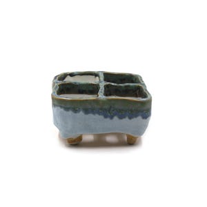 May include: A small, square ceramic planter with four compartments. The planter is glazed in shades of blue and green, with a textured finish. It has three small, rounded feet. The overall design is minimalist and modern.