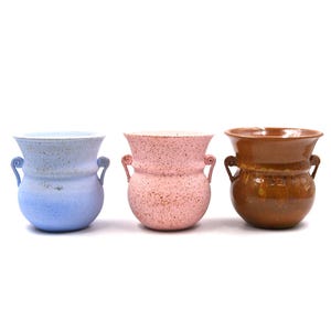 May include: Three ceramic vases in a row, each with a unique color and design. The vases are light blue, pink, and brown, with a speckled texture. Each vase has two small handles on the sides.
