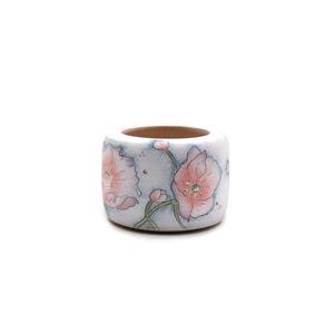 May include: A small, square ceramic container with a light blue background and pink floral designs. The container has a brown interior and is decorated with detailed illustrations of pink flowers and green stems.