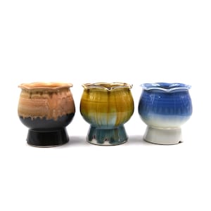 May include: Three ceramic planters with scalloped rims and pedestal bases. The left planter features a tan and black drip glaze. The center planter has a yellow, green, and blue drip glaze. The right planter displays a blue and white gradient.