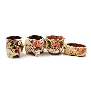 May include: Four small ceramic flower pots with floral designs. The pots are in various shapes and sizes, including round, square, and rectangular. The flowers are painted in shades of pink, yellow, and green.