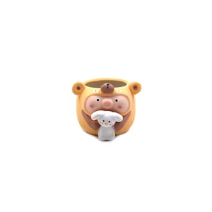 May include: A yellow ceramic planter shaped like a bear wearing a pig costume, featuring a small white rabbit figure attached to the front. The bear has brown ears and a nose, while the pig face has pink cheeks and a snout, with a white interior.