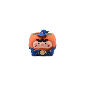 May include: A small, round ceramic planter shaped like a pig wearing a blue wizard hat and a blue robe with yellow stars. The pig has a black mustache, pink cheeks, and holds a small broom. The top portion is orange with a carved pumpkin face.