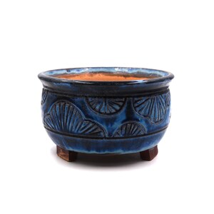 Può includere: Un vaso rotondo in ceramica blu con un motivo strutturato a forma di conchiglia all'esterno. Il vaso ha una glassa blu scuro e poggia su tre piccoli piedini in legno marrone. L'interno è di un colore marrone-arancio a contrasto.