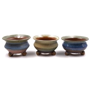 May include: Three small, ceramic bonsai pots with a rounded shape and three small feet. The pots have a brown interior and a color gradient exterior, with shades of blue, pink, and green. The pots are set against a white background.