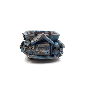 May include: A blue and brown ceramic planter shaped like a small house with a brown door and a roof. The planter is decorated with flowers and mushrooms.