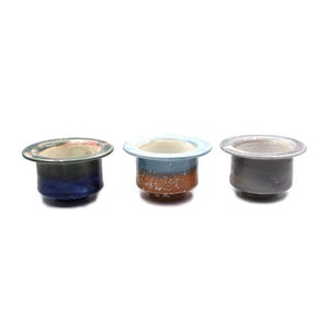 May include: Three small, handcrafted ceramic pots with a wide, flat rim. Each pot features a unique color scheme: blue and green, blue and brown, and gray. The pots are likely for small plants or decorative purposes.