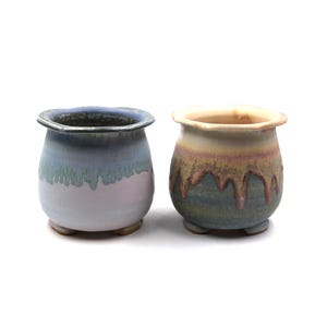 May include: Two ceramic pots with a rounded shape and small feet. One pot is light blue with a white and green glaze, the other is brown, green, and yellow. Both have a wavy rim and are suitable for small plants.