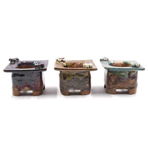 May include: Three ceramic bonsai pots with unique glaze patterns and decorative accents. The pots are square with rounded edges and have four small feet.