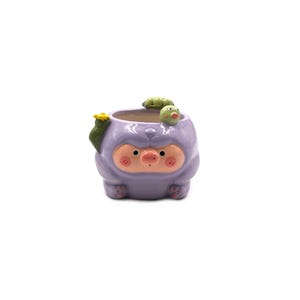 May include: A light purple ceramic planter shaped like a creature with a pink snout, rosy cheeks, and small paws. A small green caterpillar rests on the rim, and a yellow flower is attached to the side. The interior is white and hollow.