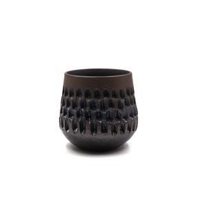 May include: A dark brown and black ceramic cup with a rounded base and a wide opening. The exterior surface features a repeating pattern of vertical, elongated indentations that create a textured, tactile appearance around the middle section.