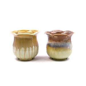 Pode incluir: Dois vasos de flores de cerâmica com borda festonada. O vaso à esquerda é marrom claro com listras brancas. O vaso à direita é um esmalte marrom e azul.
