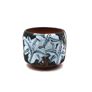 May include: A black ceramic planter with a square shape and a brown interior. The exterior features a detailed relief of light blue flowers with white accents. The planter is suitable for indoor or outdoor use.