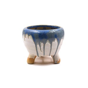 May include: A small, round ceramic planter with three short, unglazed tan feet. The exterior features a glossy glaze with a deep blue rim that drips down over a white base, creating a layered, textured appearance across the curved surface.