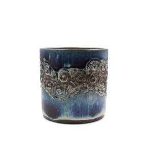 May include: A cylindrical ceramic vase with a dark blue glaze and a decorative band of swirling, raised relief patterns. The vase has a dark brown base and a lighter blue interior.