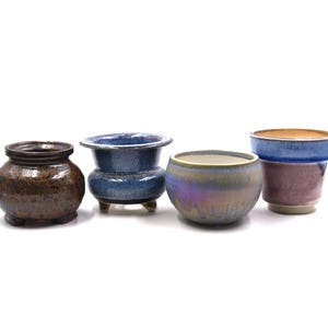 May include: Four ceramic pots in various shapes and colors. The pots include a brown, textured pot, a blue pot with three small feet, a round iridescent pot, and a purple and blue pot. These pots are suitable for plants.