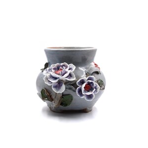 以下が含まれることがあります： 花柄の灰色の小さな陶器の花瓶。花瓶は、紫色の芯と赤いアクセントのある白い花3つで飾られています。花は盛り上がっていて、立体的に見えます。
