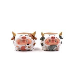 May include: Two ceramic planters shaped like cartoon cows. The left planter features brown patches and holds a small cup, while the right planter has grey patches and holds a grey whistle. Both have pink snouts, small horns, and round faces.
