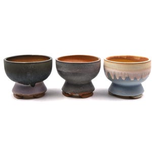 Puede incluir: Tres cuencos de cerámica hechos a mano con base de pedestal. Los cuencos tienen una variedad de colores, incluyendo verde oscuro, morado, gris y azul. El interior de los cuencos es de color naranja cálido. Sobre fondo blanco.