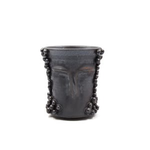 May include: A dark gray ceramic cup with a face design. The cup features closed eyes, a nose, and a mouth. Small, dark, round embellishments are arranged around the face. The cup has a smooth, slightly reflective surface.