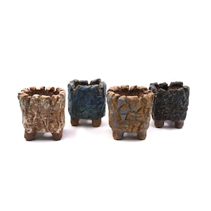 May include: Four small, square ceramic planters with textured, rough exteriors. The planters are in shades of brown, blue, and black, each with a unique, weathered appearance. They have small, rounded feet and a jagged top edge.