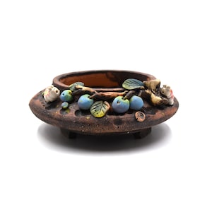 May include: A brown ceramic planter with a decorative rim featuring sculpted blue and green fruit and leaves. The planter has a low profile and sits on three small feet. The rim is decorated with small, white, sculpted figures.
