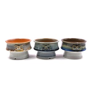 Pode incluir: Três vasos de cerâmica com base branca e parte superior envidraçada. Os vasos têm um esmalte castanho, azul e laranja com um design de laço decorativo.