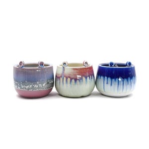May include: Three ceramic planters with unique glaze patterns. The planters are a light blue, a light green with pink accents, and a light purple with gray accents. Each planter has two small loops on the rim.