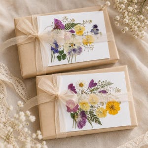 Peut inclure: Deux boîtes cadeaux rectangulaires enveloppées dans du papier kraft, chacune ornée d'une carte blanche décorée de fleurs séchées colorées. Un ruban beige transparent est noué en boucle autour de chaque boîte, fixant la carte au centre.