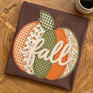 Puede incluir: Una camiseta marrón oscuro con una gran aplicación de calabaza hecha de varios parches de tela en naranja, verde y blanco con patrones de cuadros, flores, lunares y hojas. La palabra fall está bordada en letra cursiva blanca en el centro.