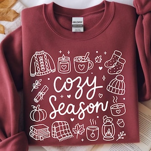 Puede incluir: Una sudadera de cuello redondo color granate con un estampado gráfico blanco con el texto "cozy Season" rodeado de ilustraciones de línea de un suéter, velas, una taza, calcetines, un gorro, una calabaza, libros, una manta y una linterna.