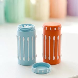 May include: Two cylindrical storage containers, one light blue and one orange, each featuring vertical white cotton swab motifs on the exterior. The blue container is open at the top, revealing white cotton swabs inside, with its matching lid placed in front.