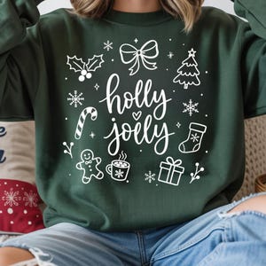 Puede incluir: Una sudadera de cuello redondo de color verde oscuro con un estampado gráfico blanco con el texto "holly jolly" en el centro. El diseño incluye ilustraciones de un lazo, un árbol de Navidad, copos de nieve, un bastón de caramelo, un calcetín, un hombre de jengibre, una taza y un regalo.