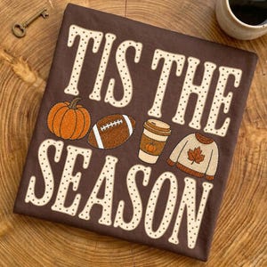 Puede incluir: Una camiseta de color marrón oscuro con el texto 'TIS THE SEASON' en letras grandes de color crema con un patrón de lunares. Entre las palabras hay gráficos bordados de una calabaza naranja, un balón de fútbol americano, un café y un suéter.