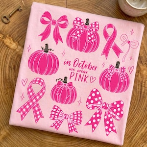 Puede incluir: Una camiseta rosa claro con un estampado gráfico de calabazas, lazos y cintas de concienciación en color rosa brillante. El texto en el centro dice "in October we wear PINK". El diseño incluye patrones de cuadros vichy, lunares y flores.