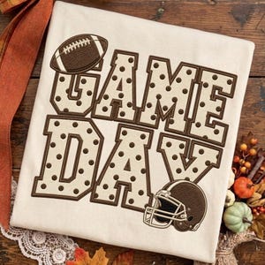 Puede incluir: Una camiseta color crema con el texto bordado GAME DAY en letras grandes con un patrón de puntos marrones. Un balón de fútbol americano marrón se sitúa sobre la palabra GAME y un casco de fútbol americano marrón bajo la palabra DAY.