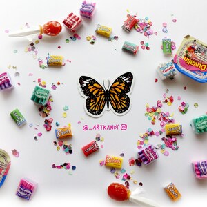 May include: A colorful butterfly sticker with orange, black, and white wings. The sticker is surrounded by colorful confetti and small packages of candy. The text "@_ARTKANDY" is written below the butterfly.