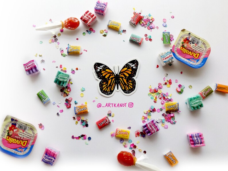 May include: A colorful butterfly sticker with orange, black, and white wings. The sticker is surrounded by colorful confetti and small packages of candy. The text "@_ARTKANDY" is written below the butterfly.