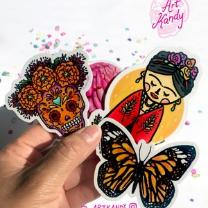May include: Three colorful stickers featuring a sugar skull with marigolds, a butterfly, and a Frida Kahlo-inspired illustration. The stickers are on a white background with confetti.