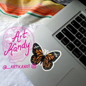 May include: A laptop computer with a butterfly sticker on the top right corner of the keyboard. The sticker is orange and black with white accents. The laptop is on a white surface with a colorful striped blanket in the background. The text "Art Kandy" and "@_ARTKANDY" is visible on the surface.