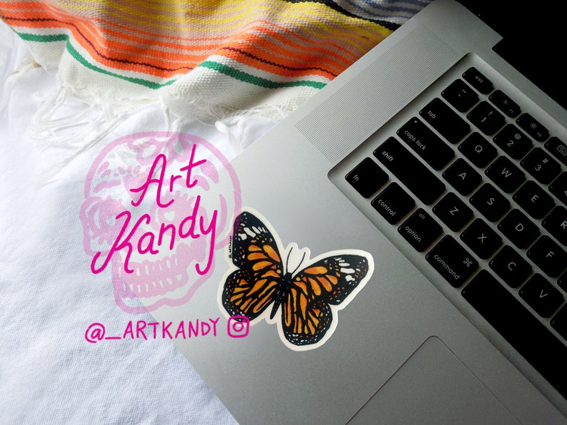 May include: A laptop computer with a butterfly sticker on the top right corner of the keyboard. The sticker is orange and black with white accents. The laptop is on a white surface with a colorful striped blanket in the background. The text "Art Kandy" and "@_ARTKANDY" is visible on the surface.