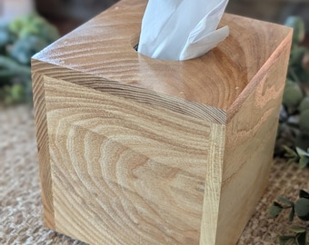 May include: A wooden tissue box with a natural wood grain finish. The box is square with a hole on top for dispensing tissues. White tissues are partially visible. The box is sitting on a woven surface.