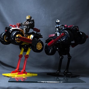 May include: Two toy action figures riding toy motorcycles with articulated legs. The figures are wearing helmets and are posed as if they are riding on a dirt track. The motorcycles are black and yellow with red accents.