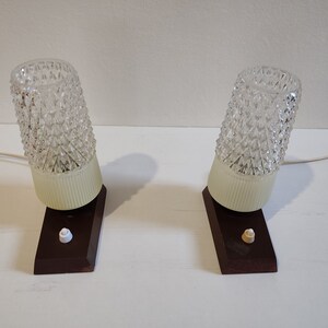 May include: Two vintage table lamps with clear glass shades featuring a diamond pattern. The lamps have a white base and a brown wooden base with a white switch.