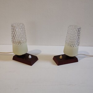 May include: Two vintage table lamps with clear glass shades and brown wooden bases. Each lamp has a white plastic shade and a small white switch.
